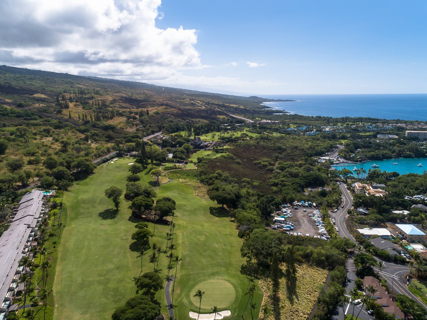 Aerial view of a lush golf course stretching toward the Pacific Ocean, surrounded by tropical greenery and mountain slopes on Hawaii's scenic coastline.