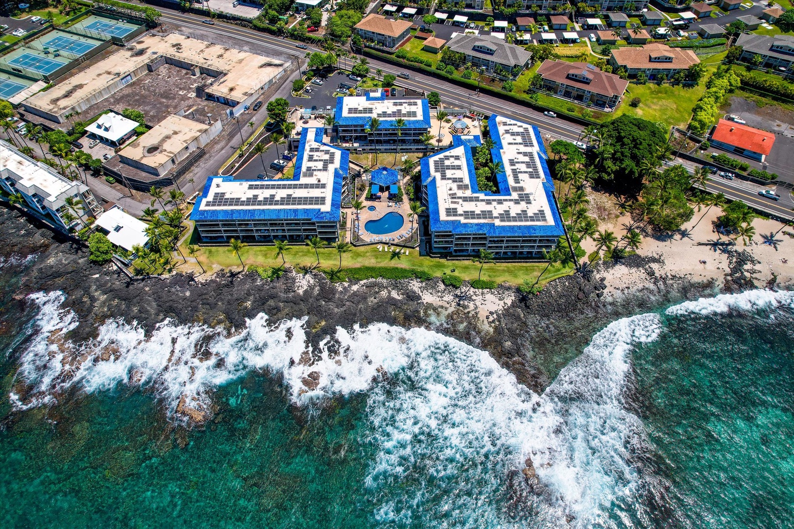 Aerial perspective showcasing the Kona Reef complex situated directly on a dramatic volcanic shoreline, where turquoise waters meet black lava rocks, surrounded by tropical landscaping and a nearby residential neighborhood.