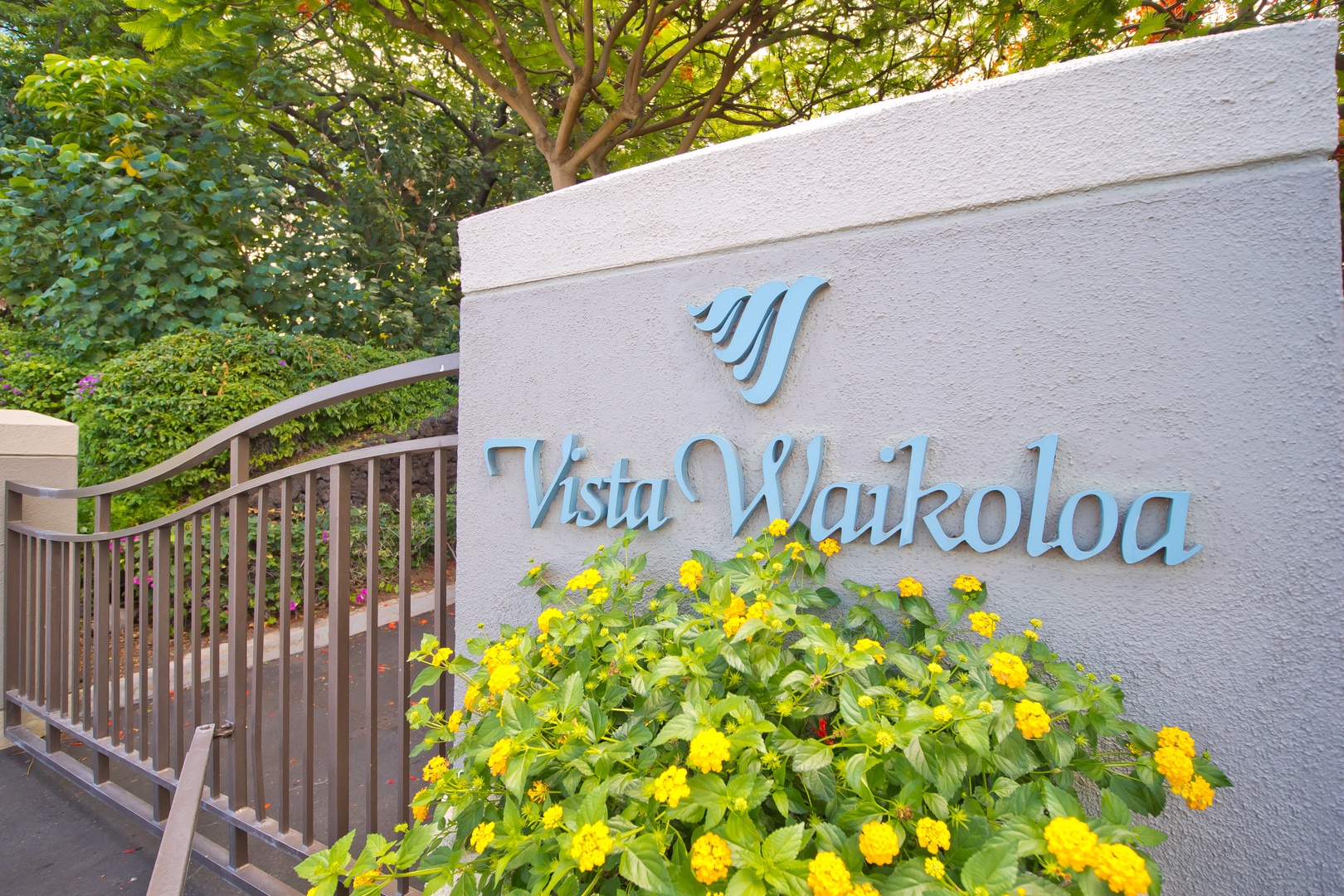 Property entrance sign featuring decorative landscaping with vibrant yellow flowers and lush tropical greenery, marking the arrival point for the vacation rental community.