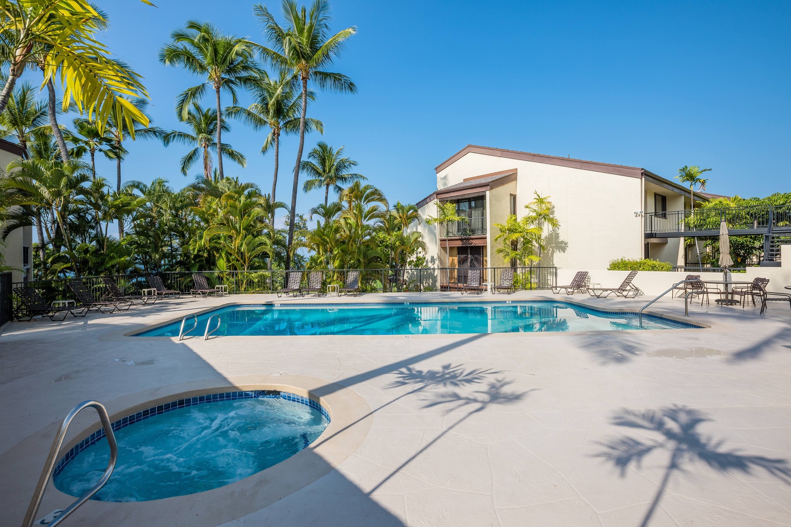 The property features a sparkling pool and hot tub surrounded by swaying palms and modern two-story buildings, all bathed in warm tropical sunshine under clear blue skies.