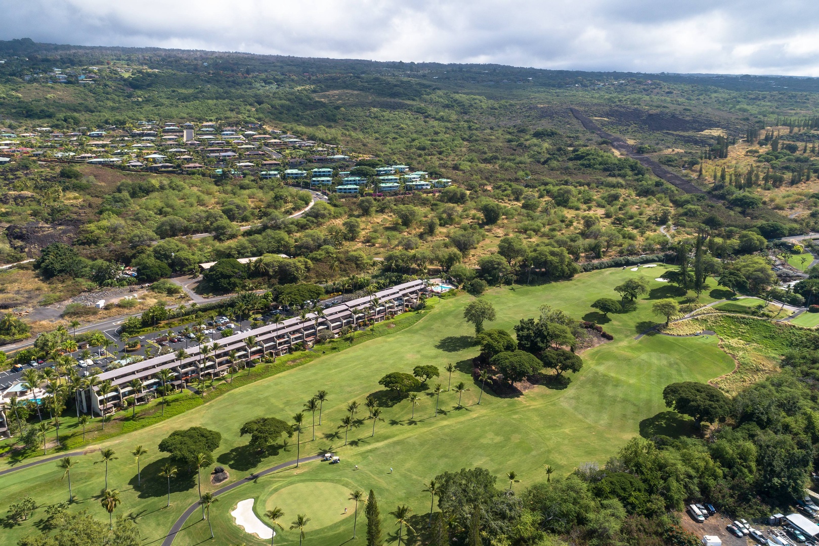 Aerial view capturing the property nestled within a lush Hawaiian landscape, bordered by manicured golf course fairways, rolling hills, and tropical vegetation.