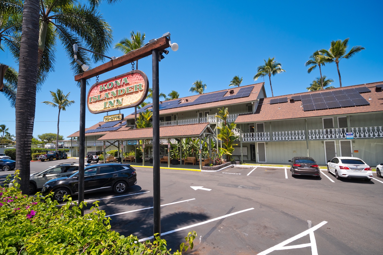 The Kona Islander Inn welcomes visitors with its distinctive signage and solar-paneled roofs, surrounded by swaying palms and convenient parking in Hawaii's sunny climate.