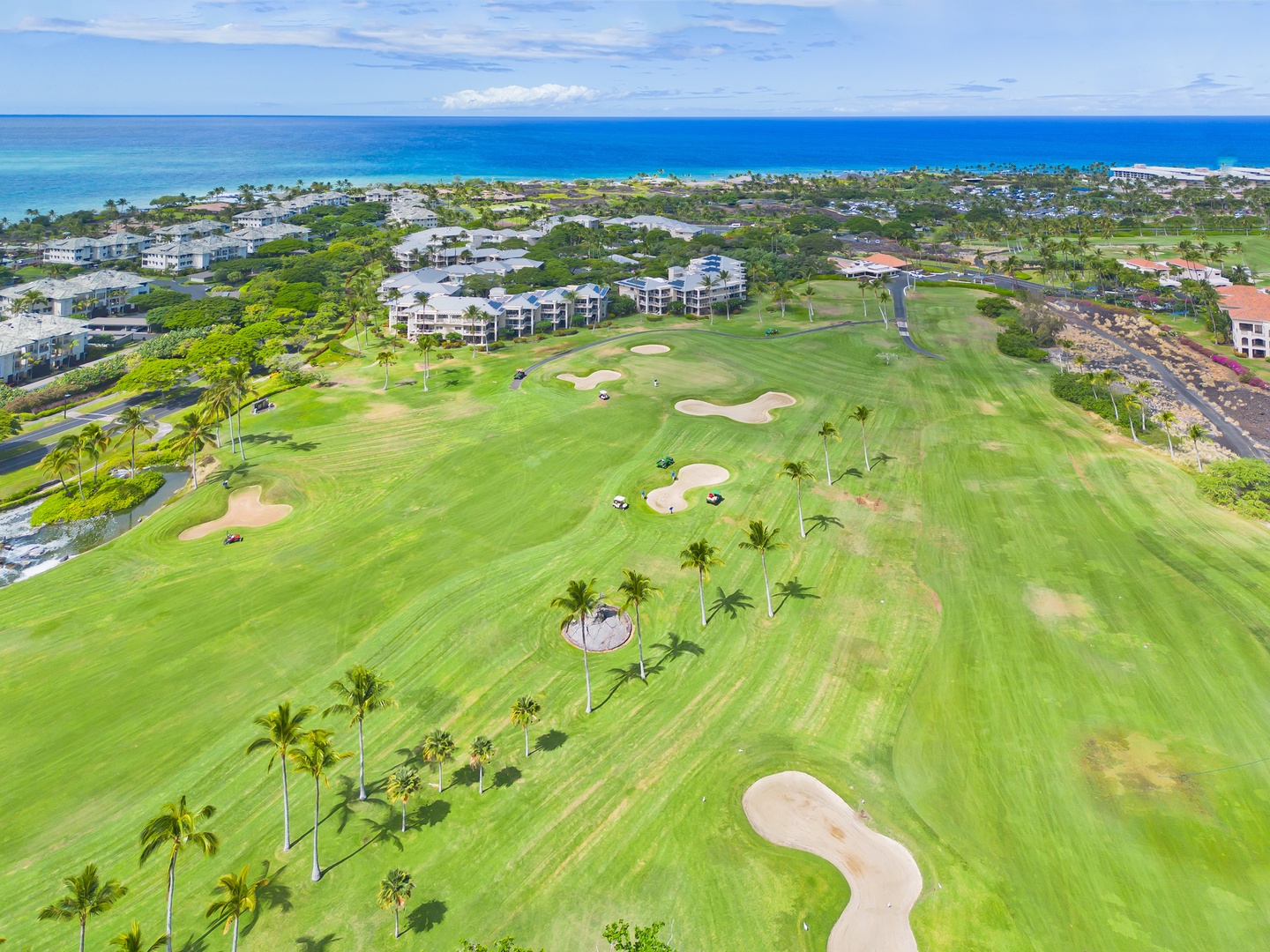 Aerial view of a pristine golf course with lush fairways, sand bunkers, and palm trees, set against a stunning ocean backdrop in a tropical coastal resort area.