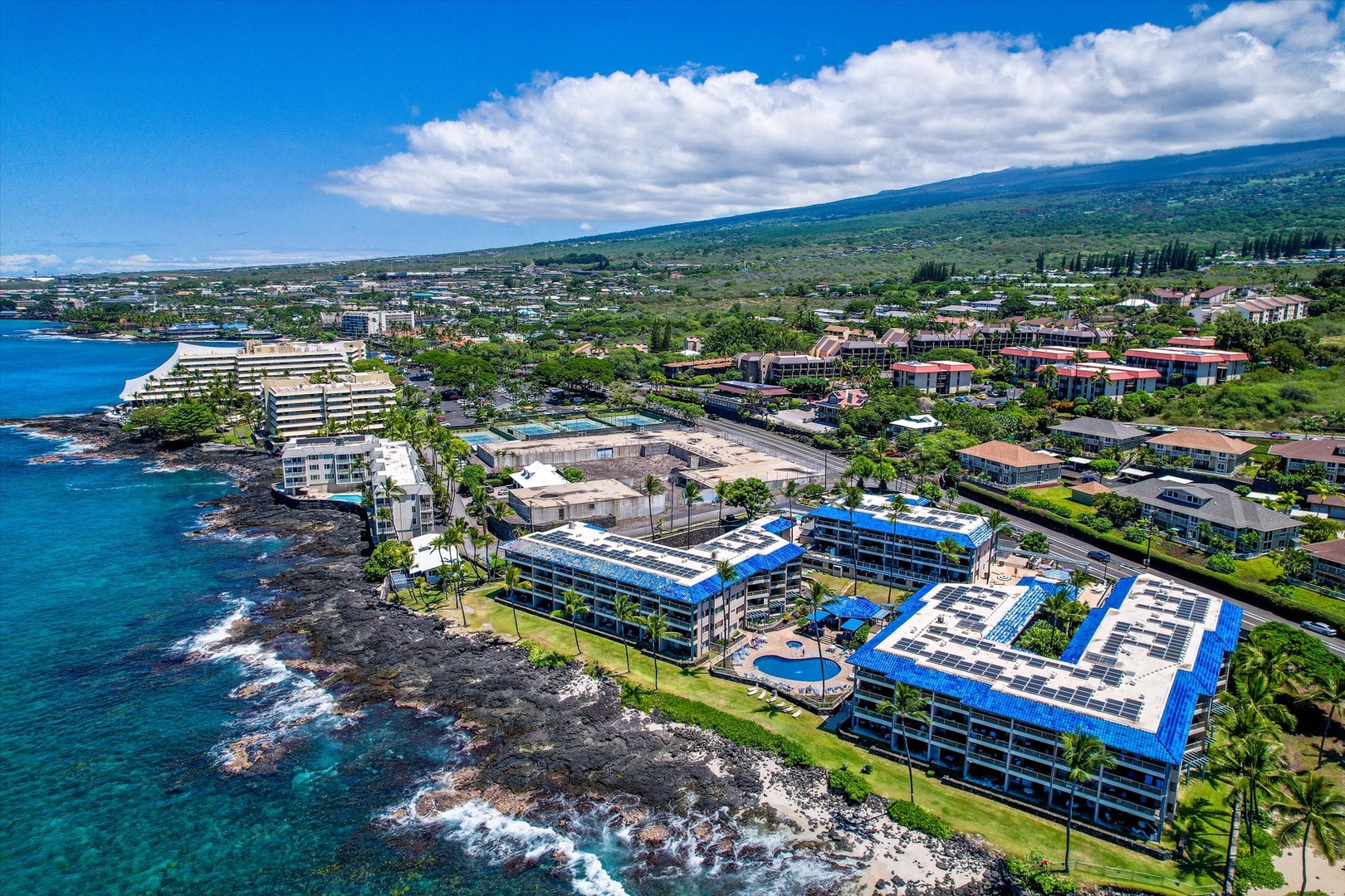 This oceanfront resort is nestled along Hawaii's dramatic volcanic coastline, surrounded by lush tropical landscape and neighboring resort areas. The property's distinctive blue roofs mark its location near the shoreline.