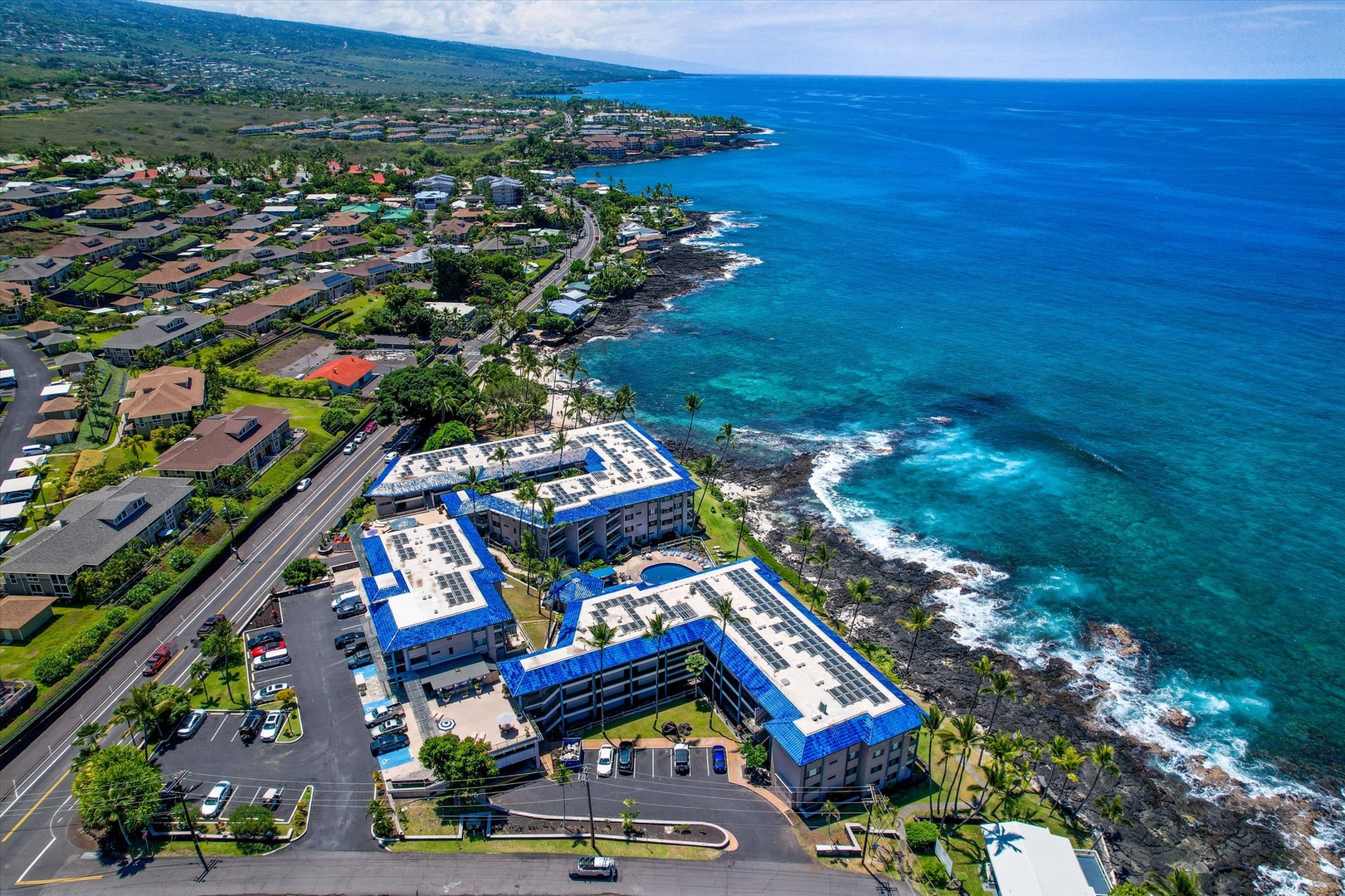 Aerial view of the beachfront property complex nestled along Hawaii's dramatic volcanic coastline, with turquoise waters meeting rugged lava rock shores and mountain vistas beyond.