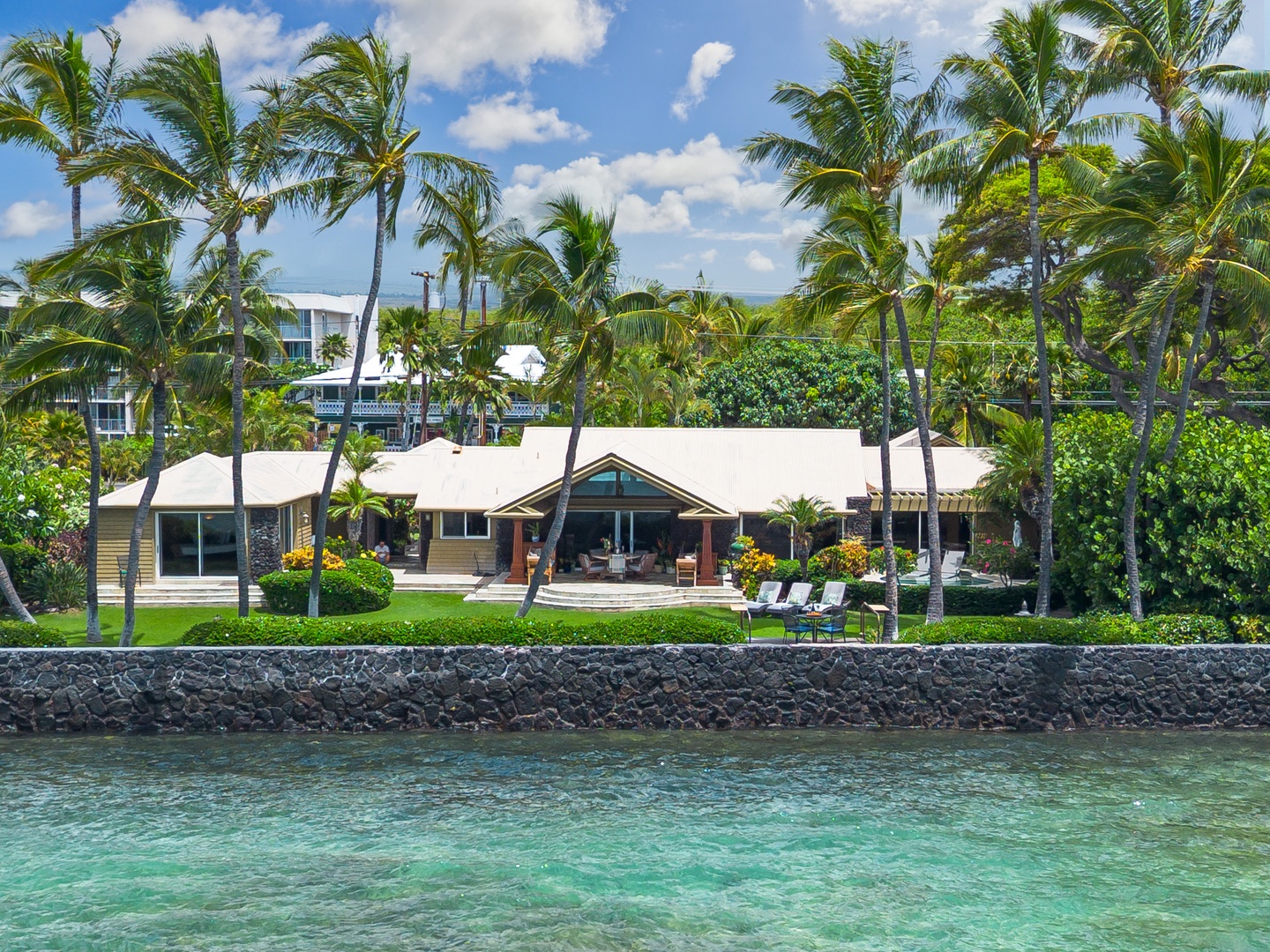 A waterfront estate framed by swaying palms and manicured tropical landscaping, set along crystalline turquoise waters with a volcanic stone seawall.