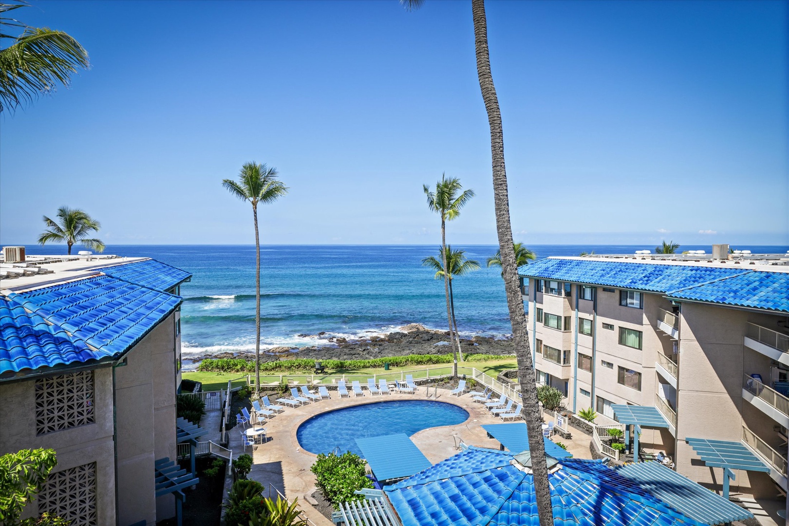 Lanai view showcasing the property's oceanfront location with a poolside courtyard, framed by tropical palms and blue tile roofs overlooking the Pacific shoreline.