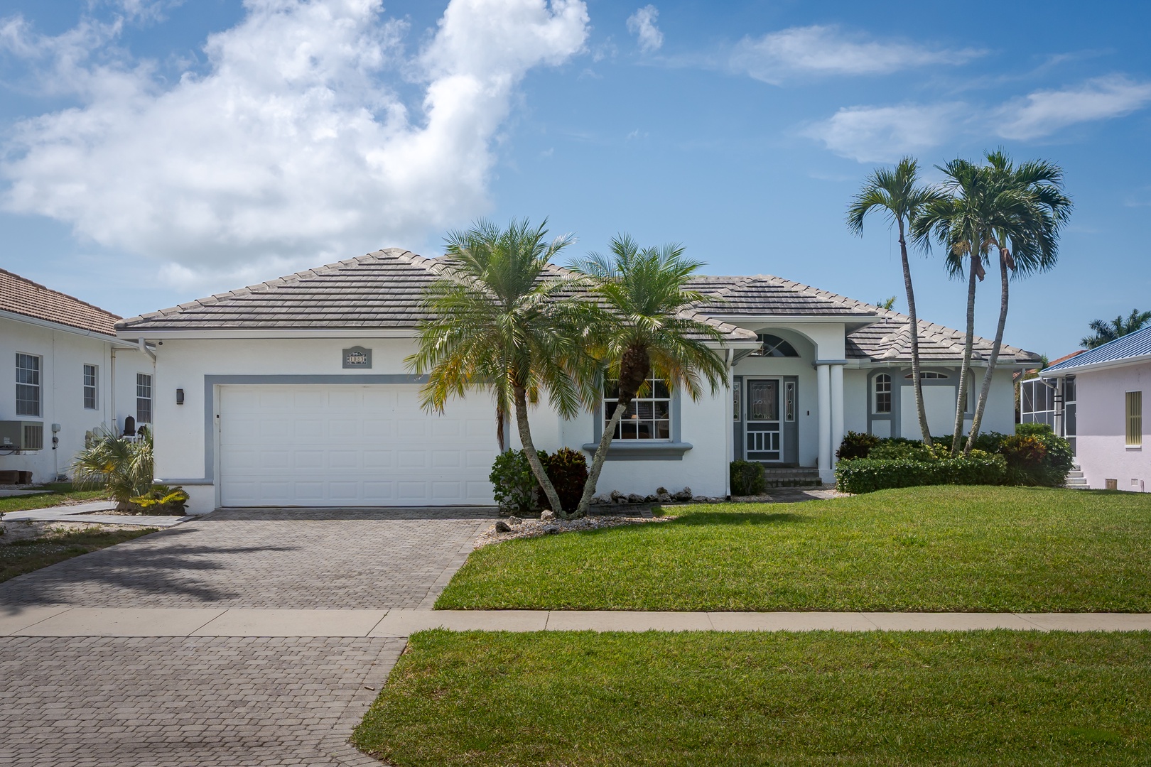 This elegant single-story vacation home features tropical landscaping and a spacious two-car garage, nestled in a peaceful residential neighborhood under sunny skies.