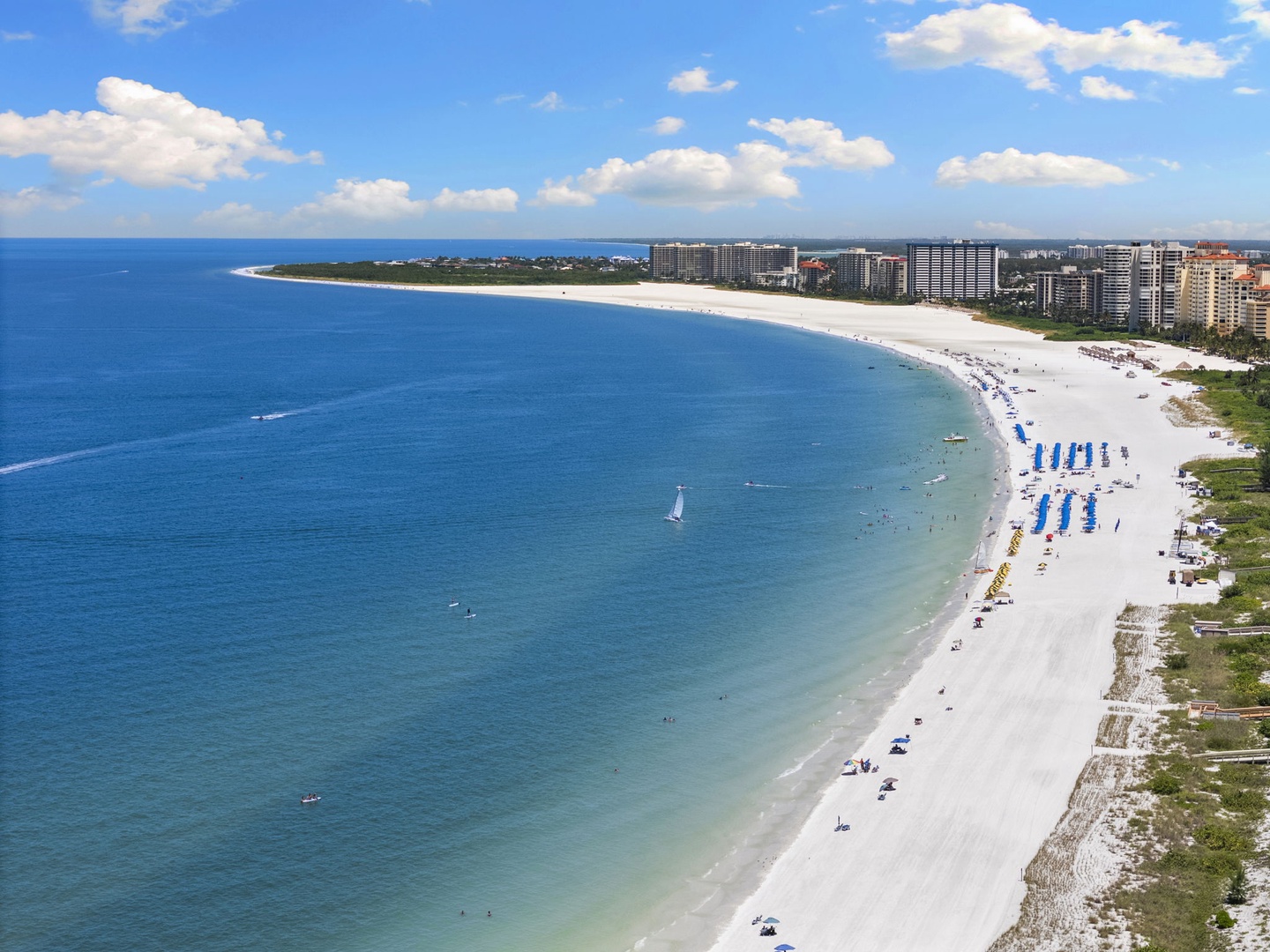 An expansive white sand beach stretches along turquoise waters, with coastal development visible in the background and the Gulf waters extending to the horizon.