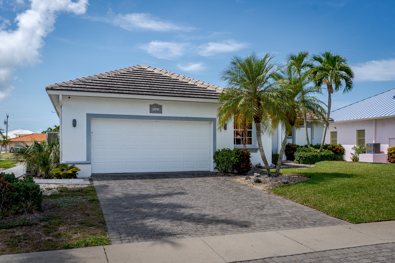 A charming single-story home with a tile roof and two-car garage, framed by tropical palm trees in a peaceful residential neighborhood under sunny blue skies.
