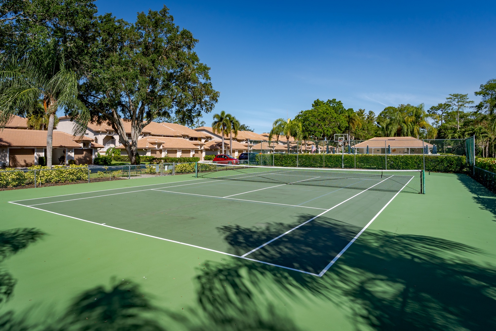 The community features a private tennis court surrounded by lush tropical landscaping and residential buildings with terracotta roofs.