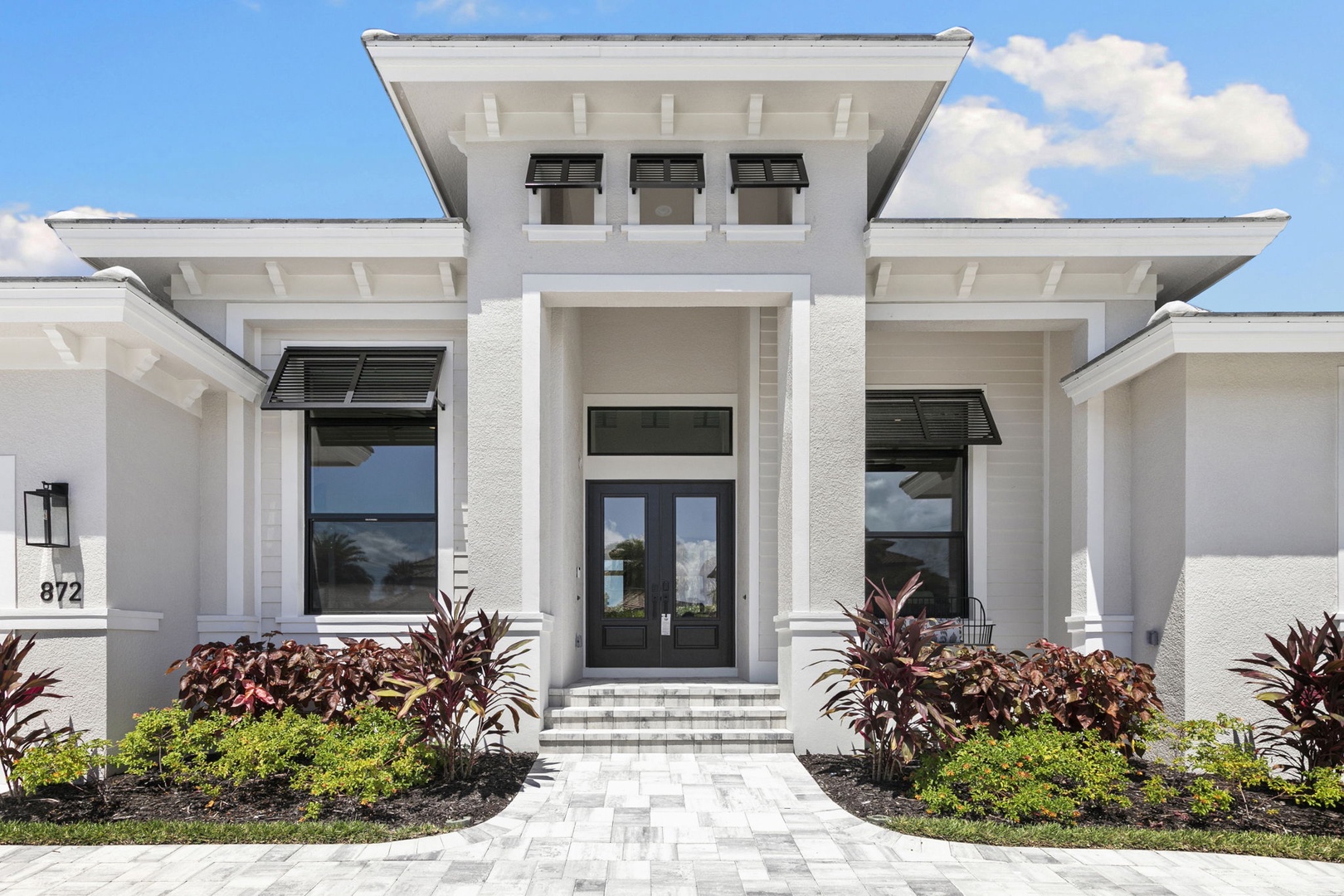 Modern tropical architecture welcomes you with clean white lines, a striking double-door entrance, and lush landscaping. The elegant paver walkway and contemporary design create a sophisticated arrival experience.
