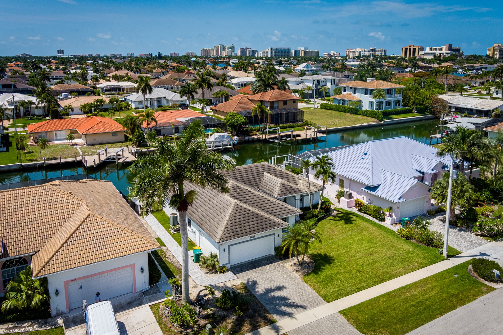 Aerial view of a waterfront neighborhood with palm-lined streets, canal-front homes with private docks, and the city skyline visible in the distance.