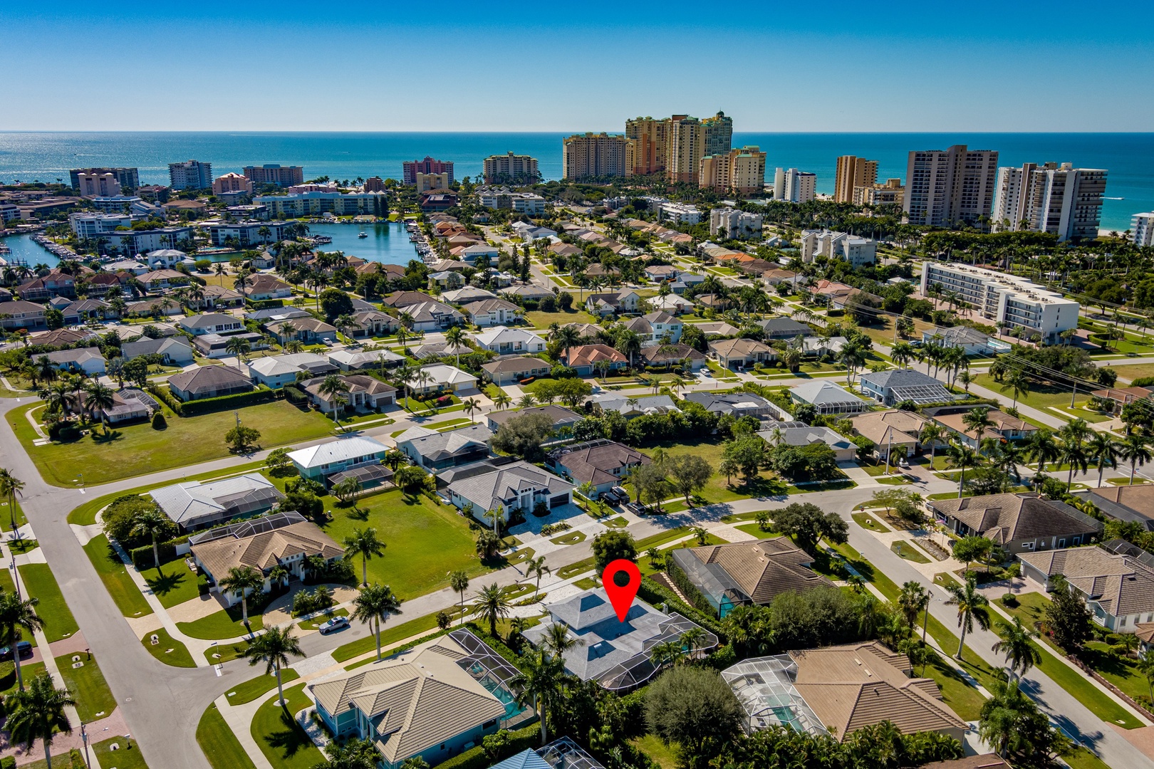 Aerial view of a coastal neighborhood with residential homes, palm trees, and beachfront high-rises in the distance.