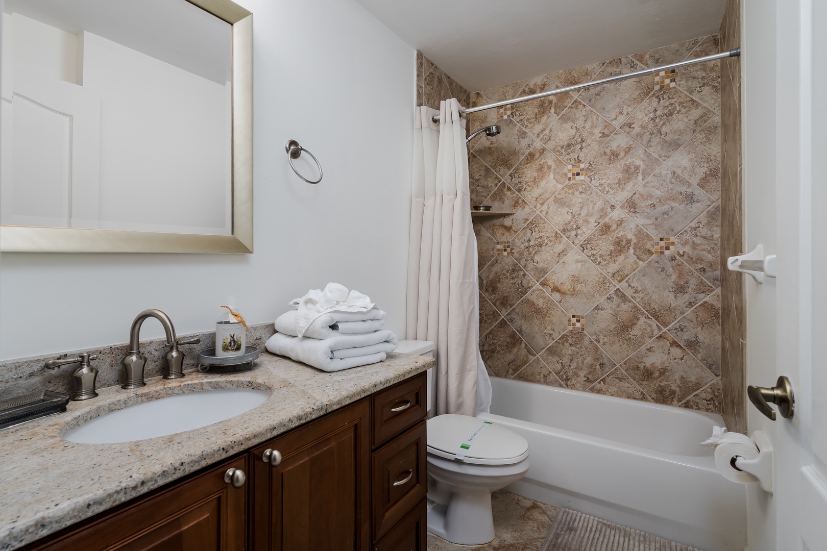 Step into your private bathroom featuring elegant granite countertops, warm wood cabinetry, and a beautifully tiled shower-tub combination. Fresh towels await your arrival.