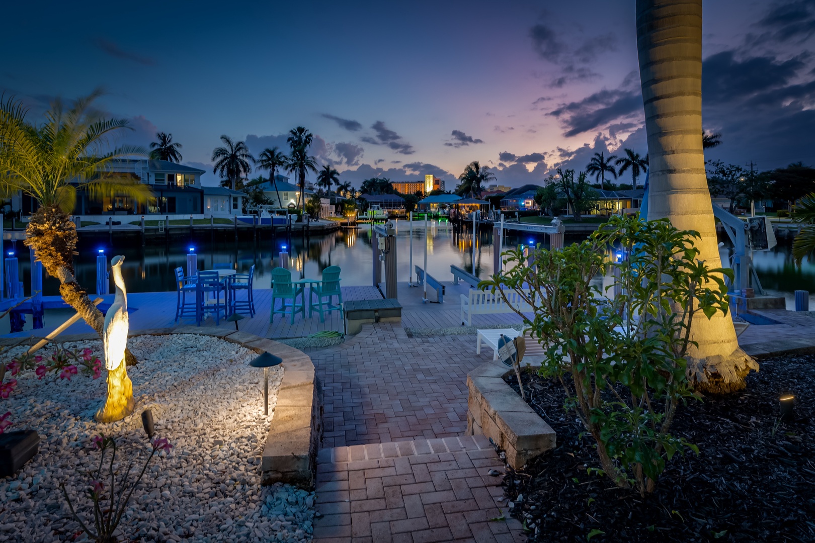 A waterfront community showcases illuminated homes along tranquil canals, with palm trees and colorful outdoor seating framing a scenic dock area at twilight.