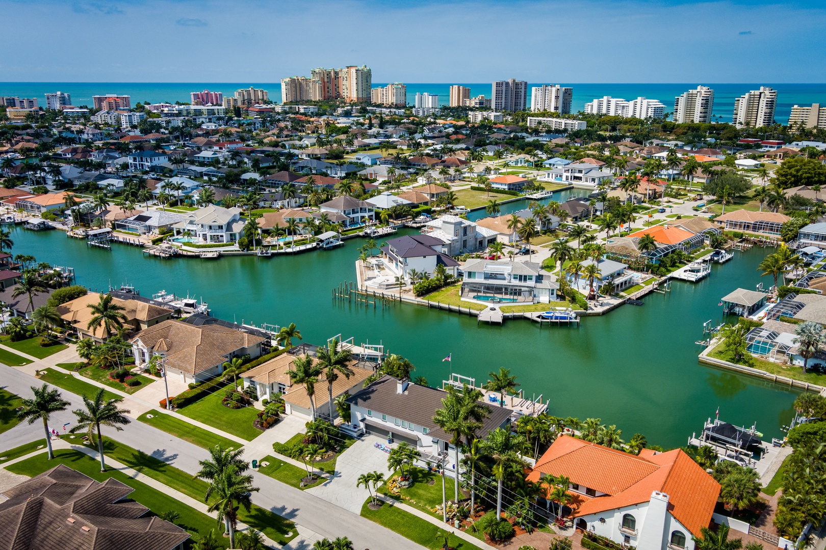 Aerial view of a waterfront neighborhood with canal-access homes, palm-lined streets, and beach high-rises in the distance, showcasing the property's coastal setting.
