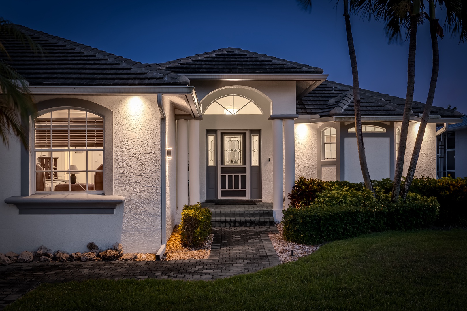 Elegant single-story home with lush tropical palms and beautifully lit arched entrance at twilight. The property features a brick paver walkway and manicured landscaping.
