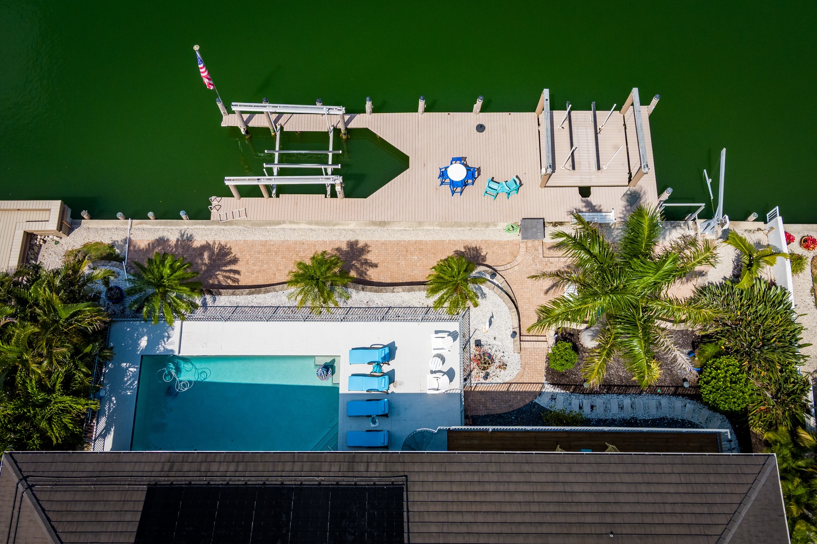 Aerial view of a waterfront vacation rental featuring a swimming pool, private dock extending into emerald waters, and lush tropical landscaping with palm trees throughout the property.