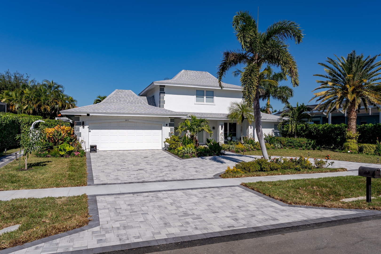 Modern white vacation home with tropical landscaping and paver driveway in sunny Florida neighborhood.