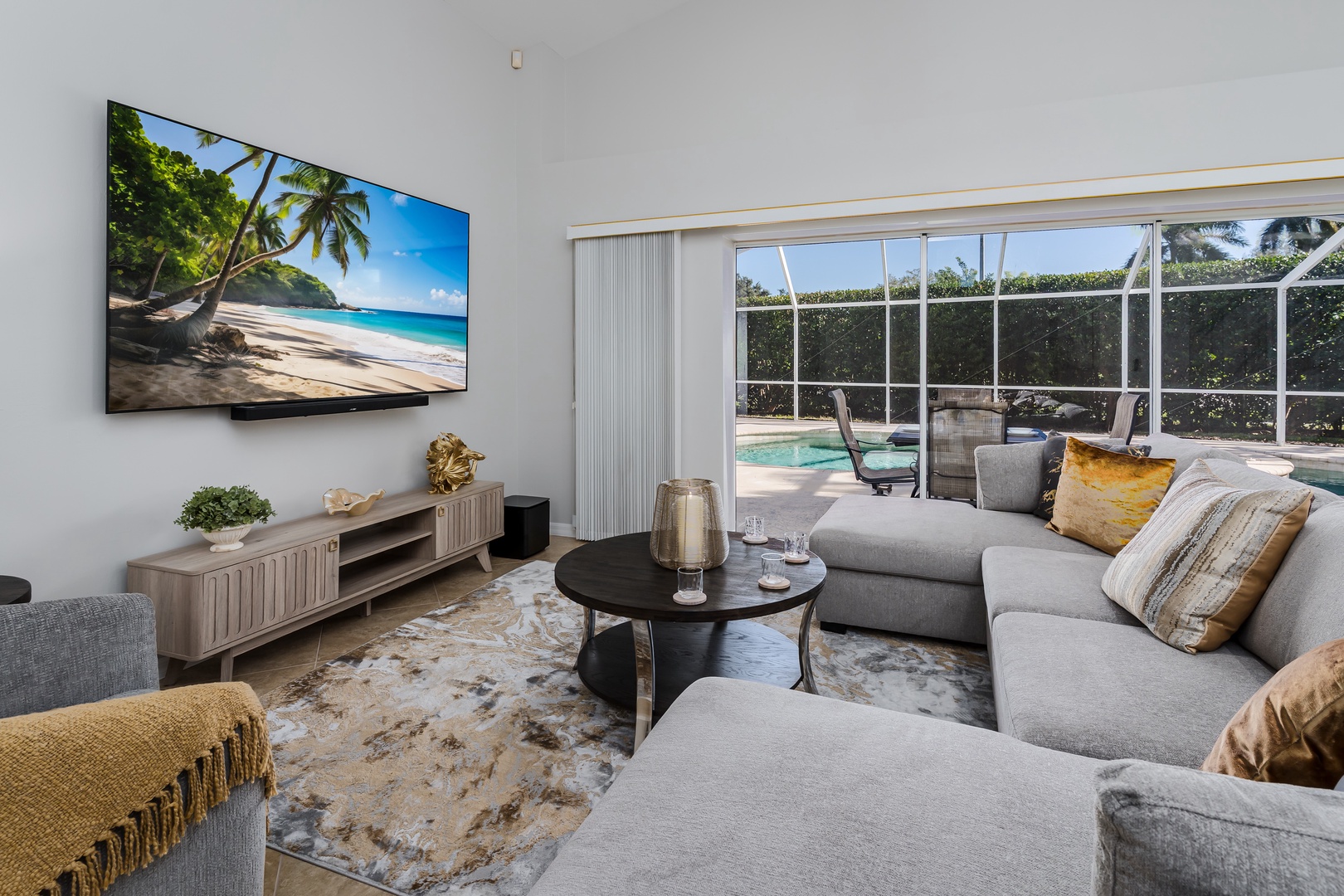 Relax in your spacious living room with plush gray sectional, large TV, and glass doors opening to your private pool area.