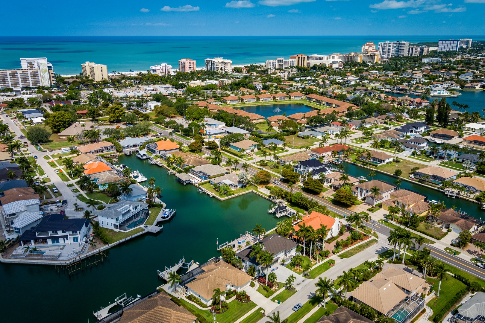 Aerial view of a vibrant coastal neighborhood featuring waterfront homes with private boat docks along calm turquoise canals, positioned between serene residential streets and pristine beachfront high-rises.
