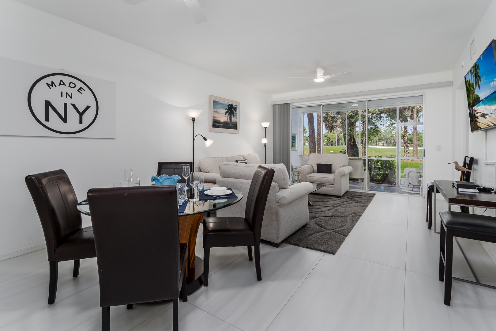 Step into your bright, open living space where sleek modern furnishings meet relaxed comfort. Floor-to-ceiling glass doors frame tranquil views of palm trees and lush greenery, inviting you to unwind in style.