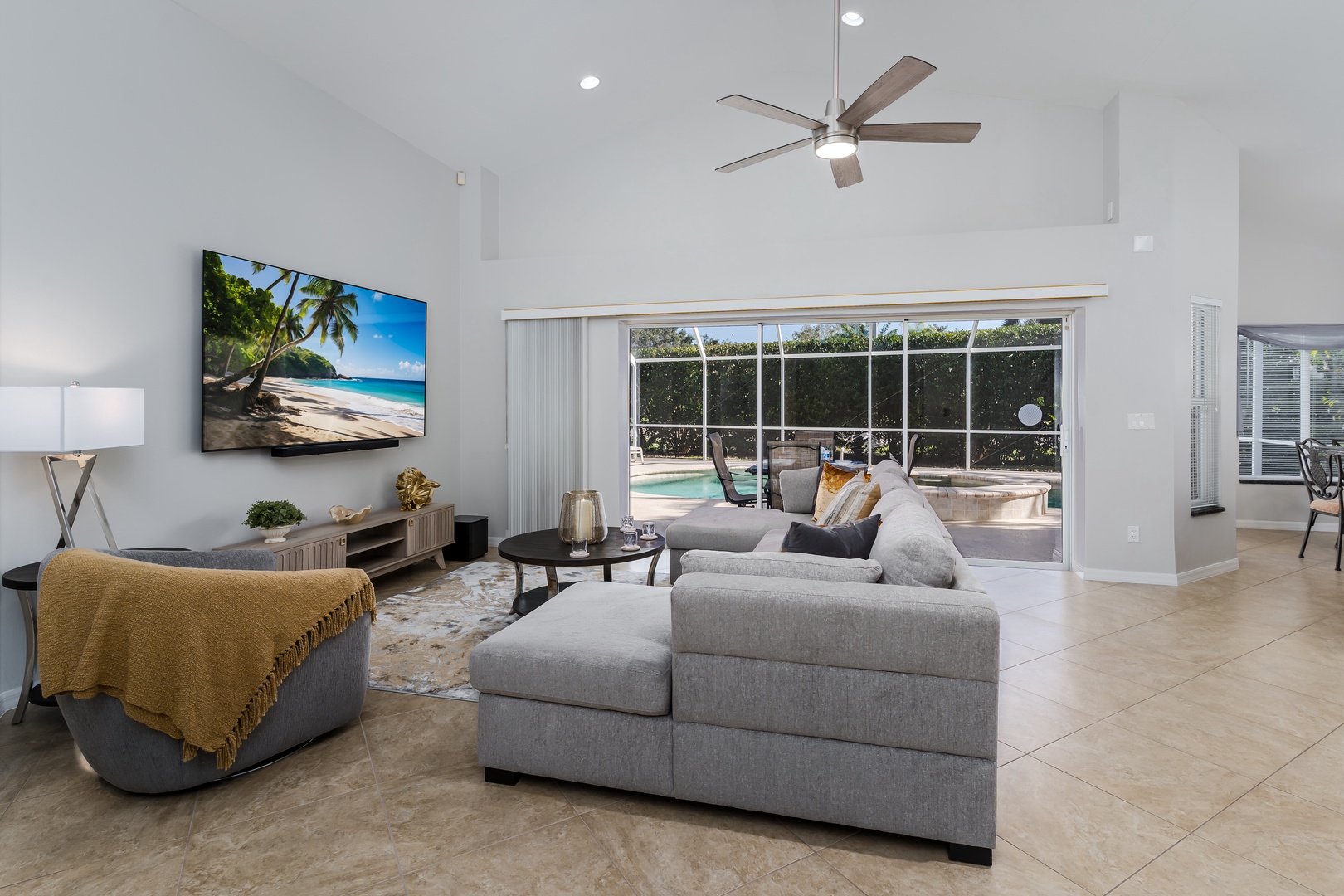 Relax in your bright living room with comfortable gray sectional, large TV, and stunning pool views through floor-to-ceiling windows.