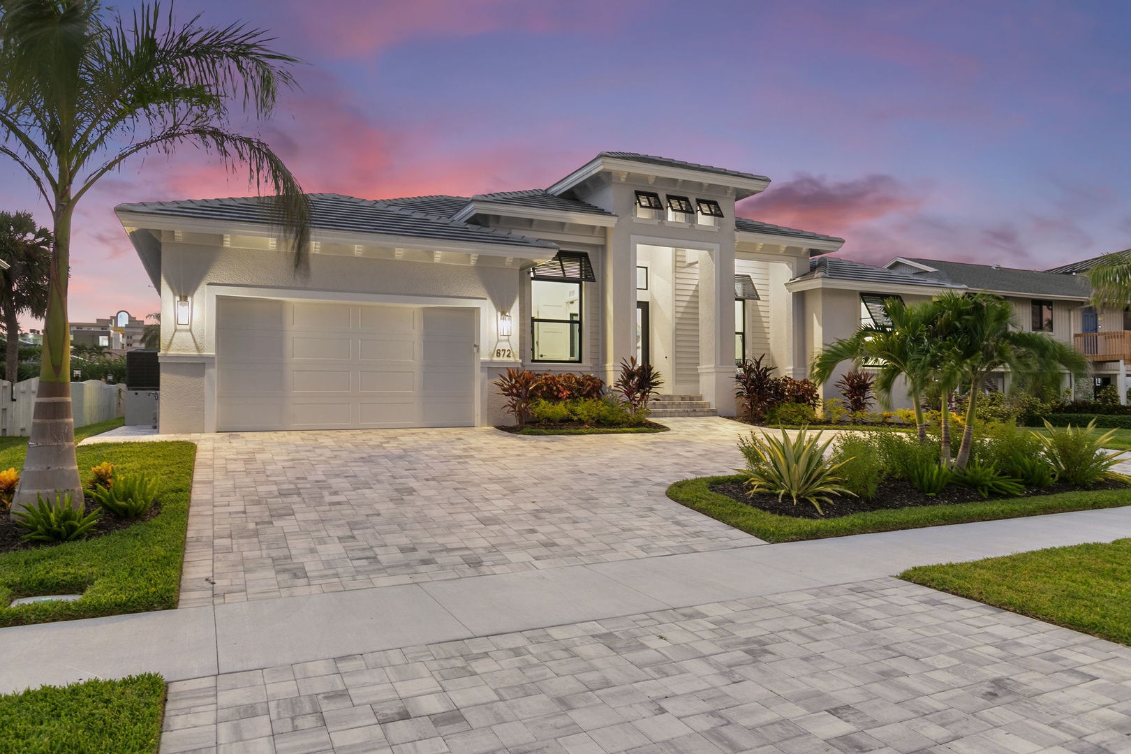 A stunning contemporary home welcomes guests at sunset, surrounded by lush tropical landscaping and elegant paver driveway in a peaceful residential neighborhood.