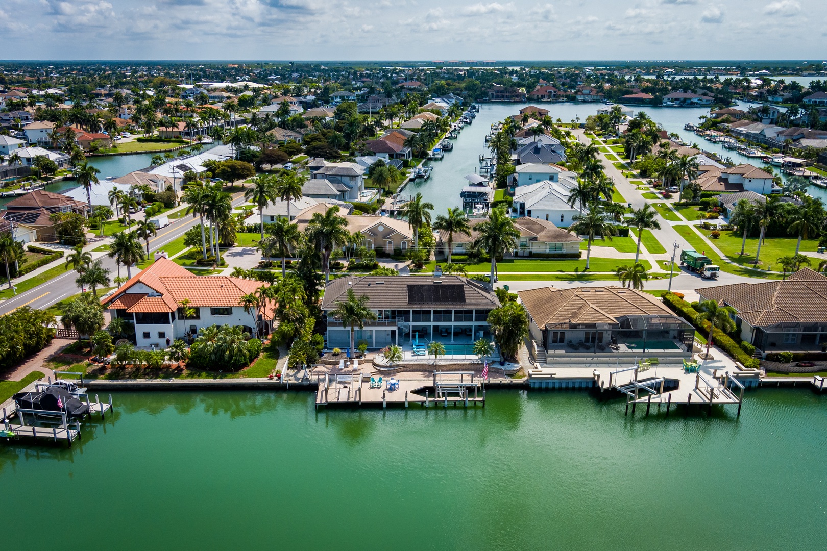 Waterfront neighborhood featuring canal-side homes with private boat docks, surrounded by lush palm trees and calm turquoise waters stretching to the ocean horizon.