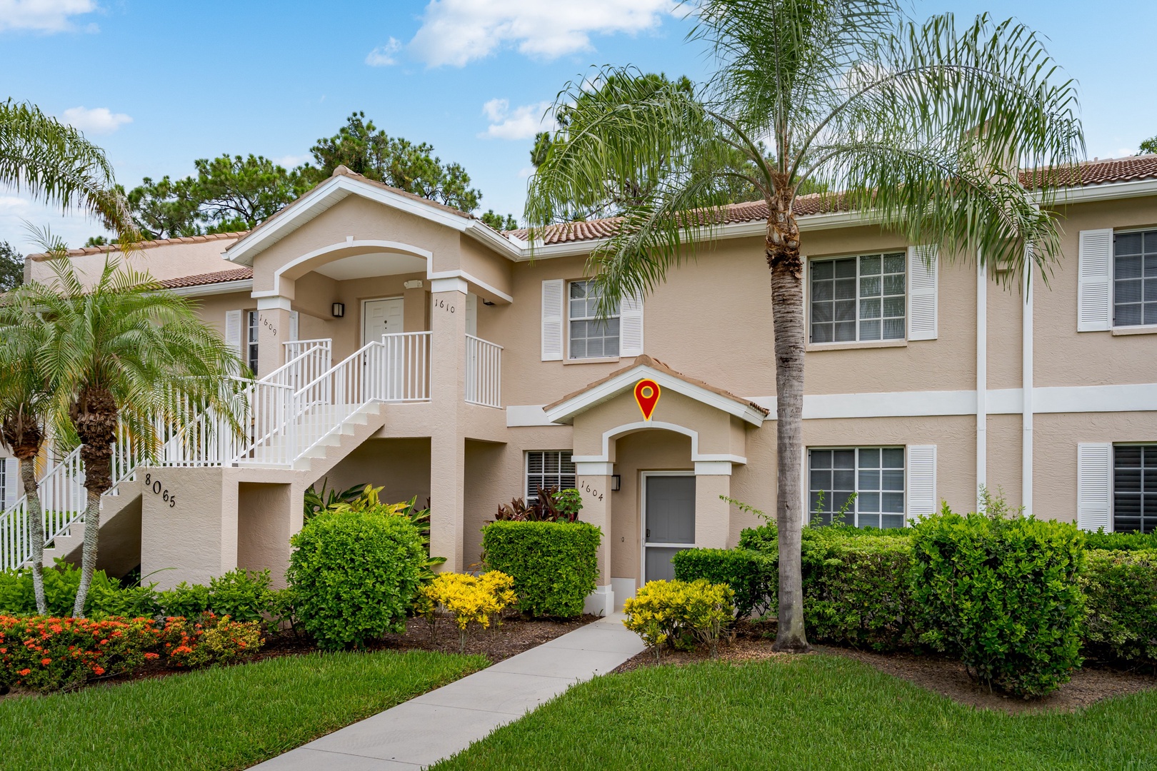 A welcoming beige stucco property with elegant arched entryways, framed by swaying palms and lush tropical landscaping beneath a bright blue sky.