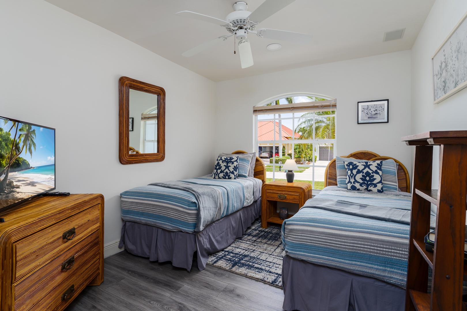Your coastal-inspired bedroom offers two inviting twin beds with fresh linens, a ceiling fan, and a bright window framing tropical palms and sunny Florida skies beyond.