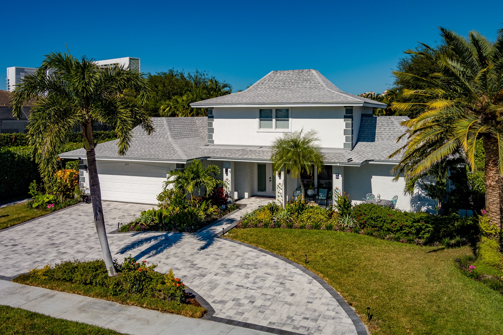 Modern tropical home with pristine landscaping and curved paver driveway, nestled among swaying palms in a quiet residential setting.