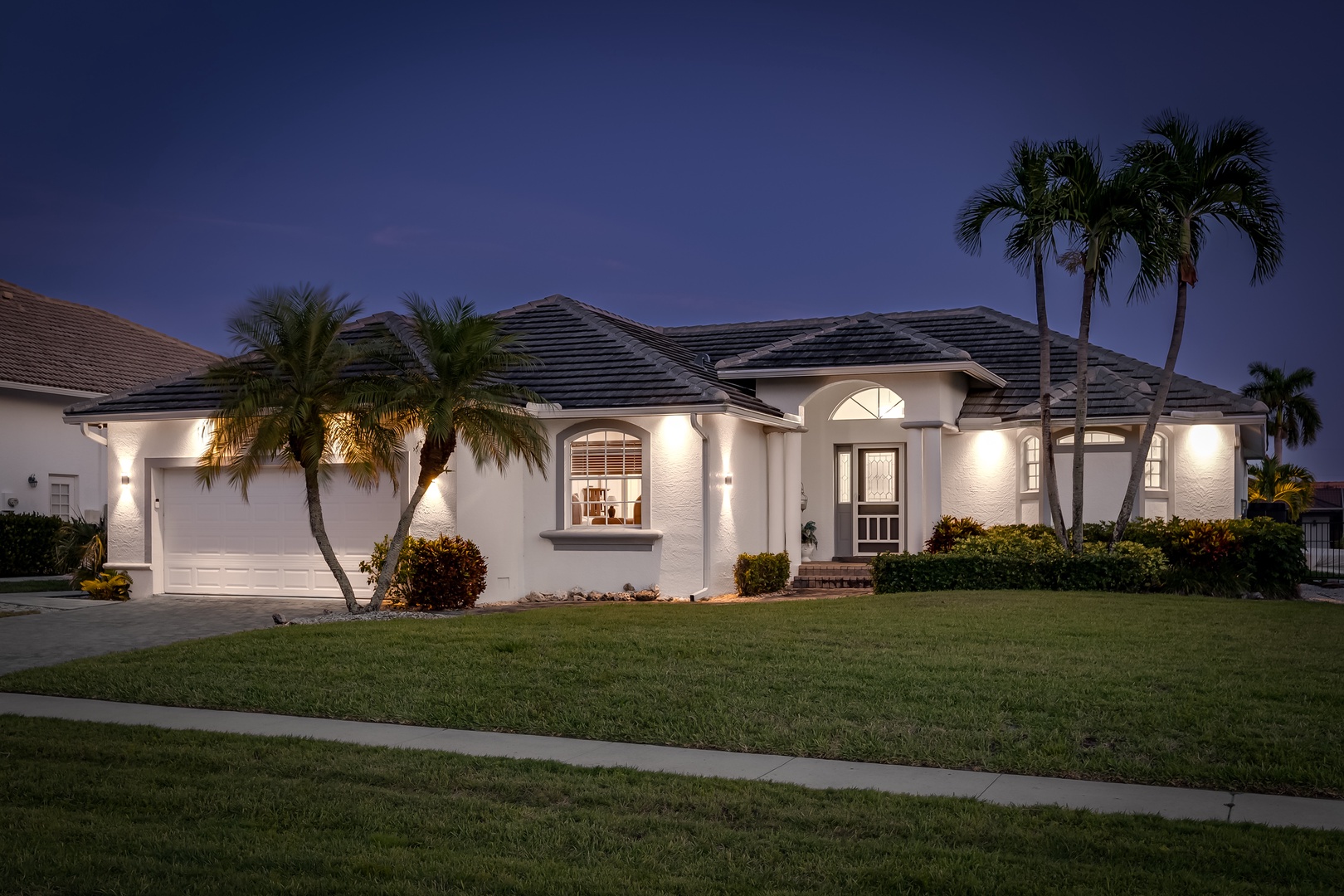 Elegant single-story home features classic architecture, palm-lined entrance, and warm twilight lighting in a peaceful residential setting.