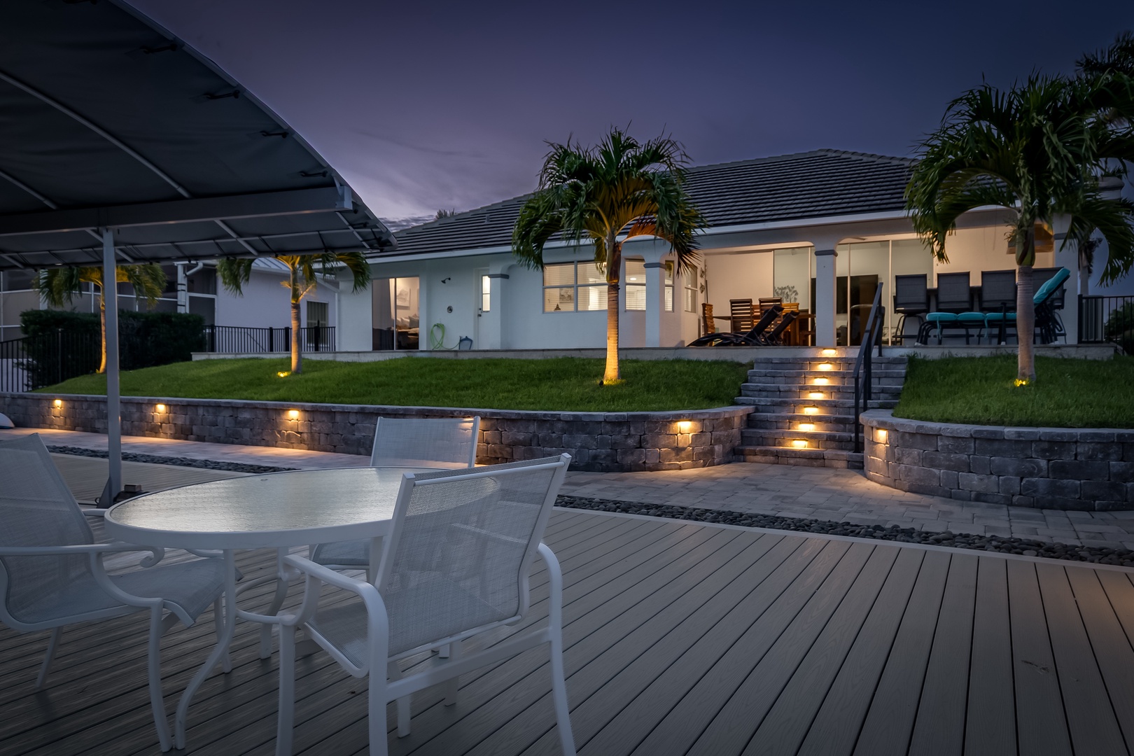 A tropical retreat bathed in twilight glow, where warm accent lighting guides the way through tiered landscaping. Palm trees stand sentinel while the open interior invites relaxation and resort-style living.