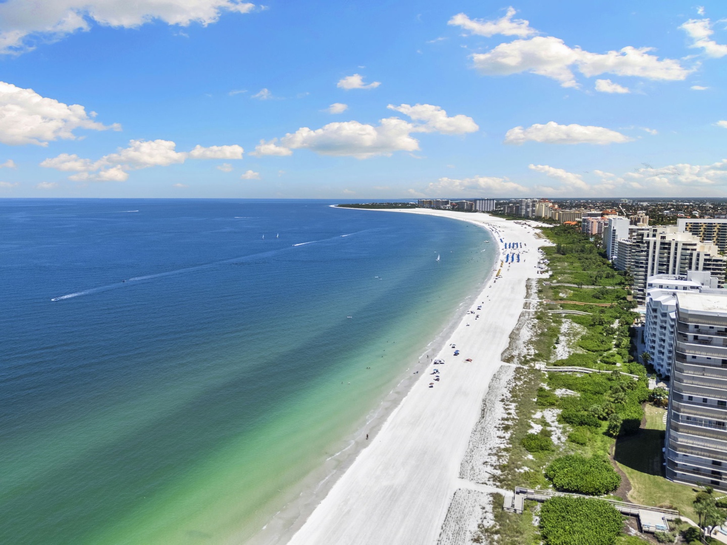 An aerial view captures the pristine white-sand beach stretching along the turquoise waters, with resort buildings nestled among green spaces along the shore—a stunning backdrop to this coastal destination.