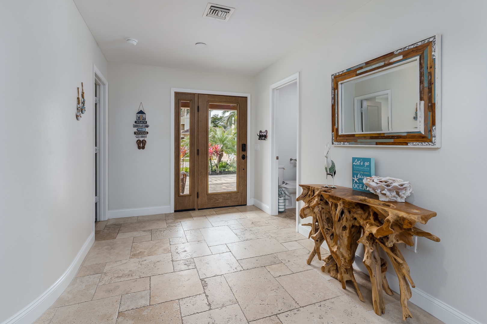Step into your bright entryway featuring unique driftwood furniture and coastal charm, where French doors reveal tropical paradise awaiting outside.