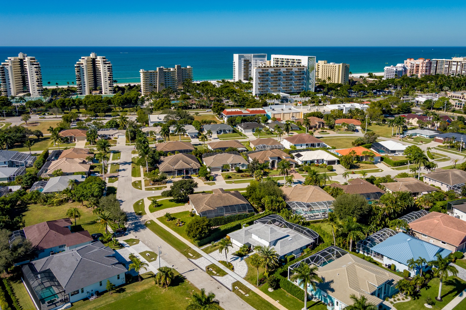 Aerial view showcases a coastal neighborhood with residential homes, tree-lined streets, and beachfront high-rises in the distance.