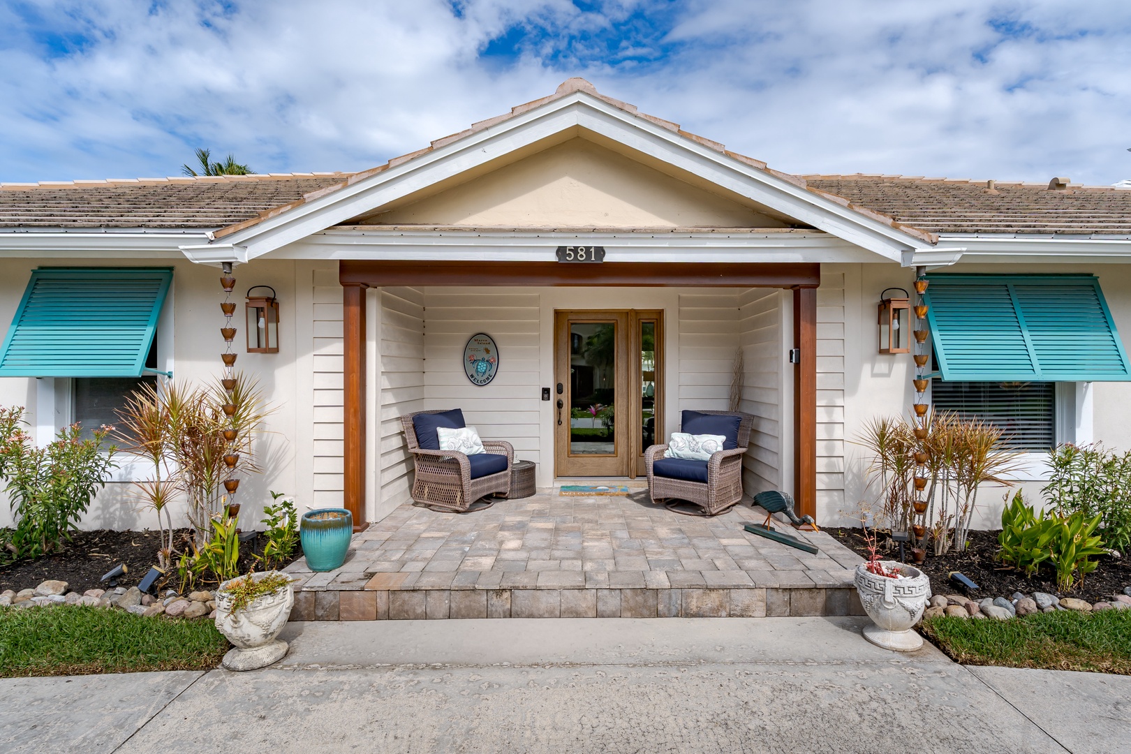 Charming coastal cottage entrance with turquoise shutters and welcoming front porch featuring comfortable seating under blue skies.
