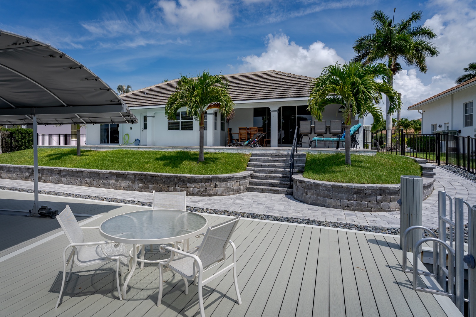 A spacious waterfront property in a tropical neighborhood, featuring tiered landscaping with palm trees, a covered patio with outdoor seating, and a private dock with dining area overlooking the canal.