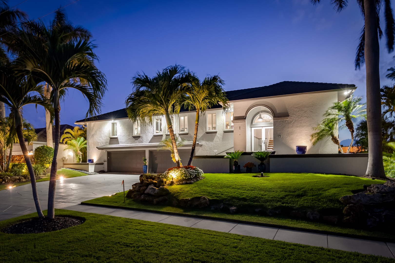 An elegant two-story vacation home featuring Mediterranean architecture, surrounded by palm trees and professionally lit landscaping with a welcoming arched entrance.