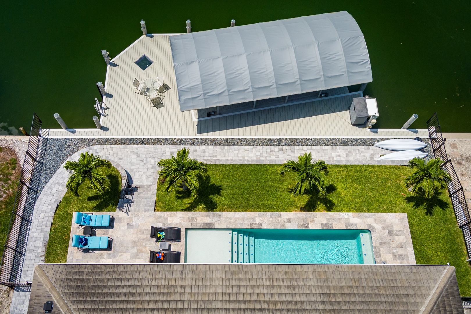 Aerial view showcasing the waterfront property with swimming pool, covered dock, lawn area with palm trees, and direct access to the water—part of the surrounding amenities.