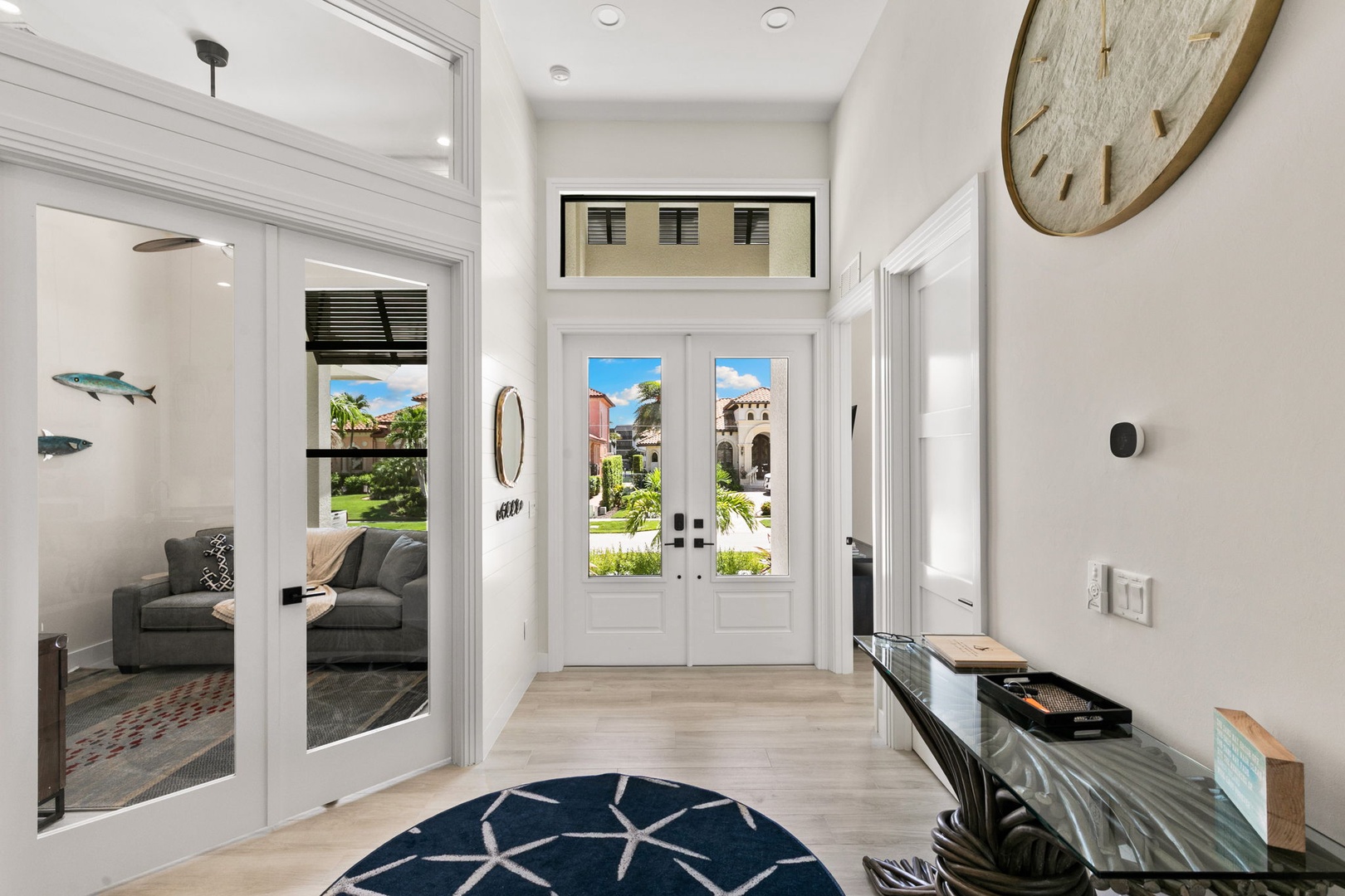 Step into an airy, light-filled foyer with soaring ceilings and coastal charm. Sunlight pours through double doors revealing lush gardens outside, while the adjacent living room invites you to unwind in style.