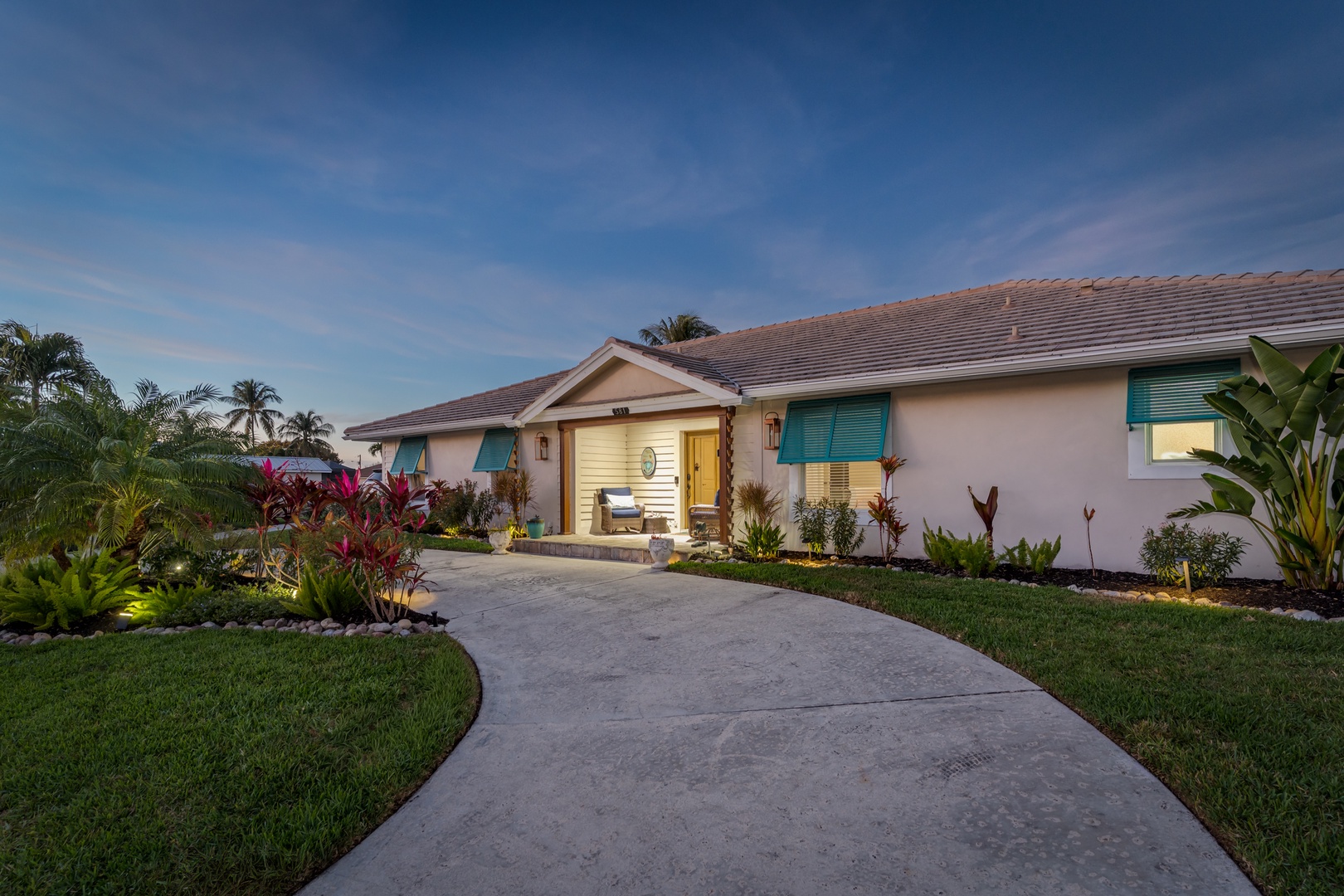 A tropical paradise awaits with this charming villa featuring vibrant landscaping, palm trees, and turquoise shutters under endless blue skies.
