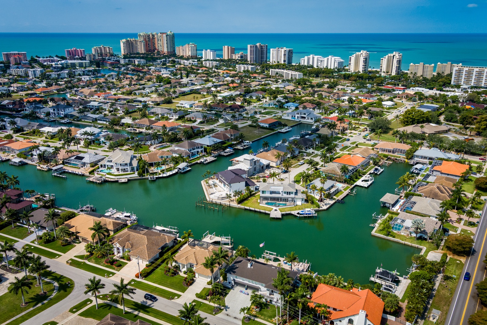 Aerial view of coastal community with waterfront homes along calm canals, distant beachfront high-rises, and tropical landscaping throughout the neighborhood.