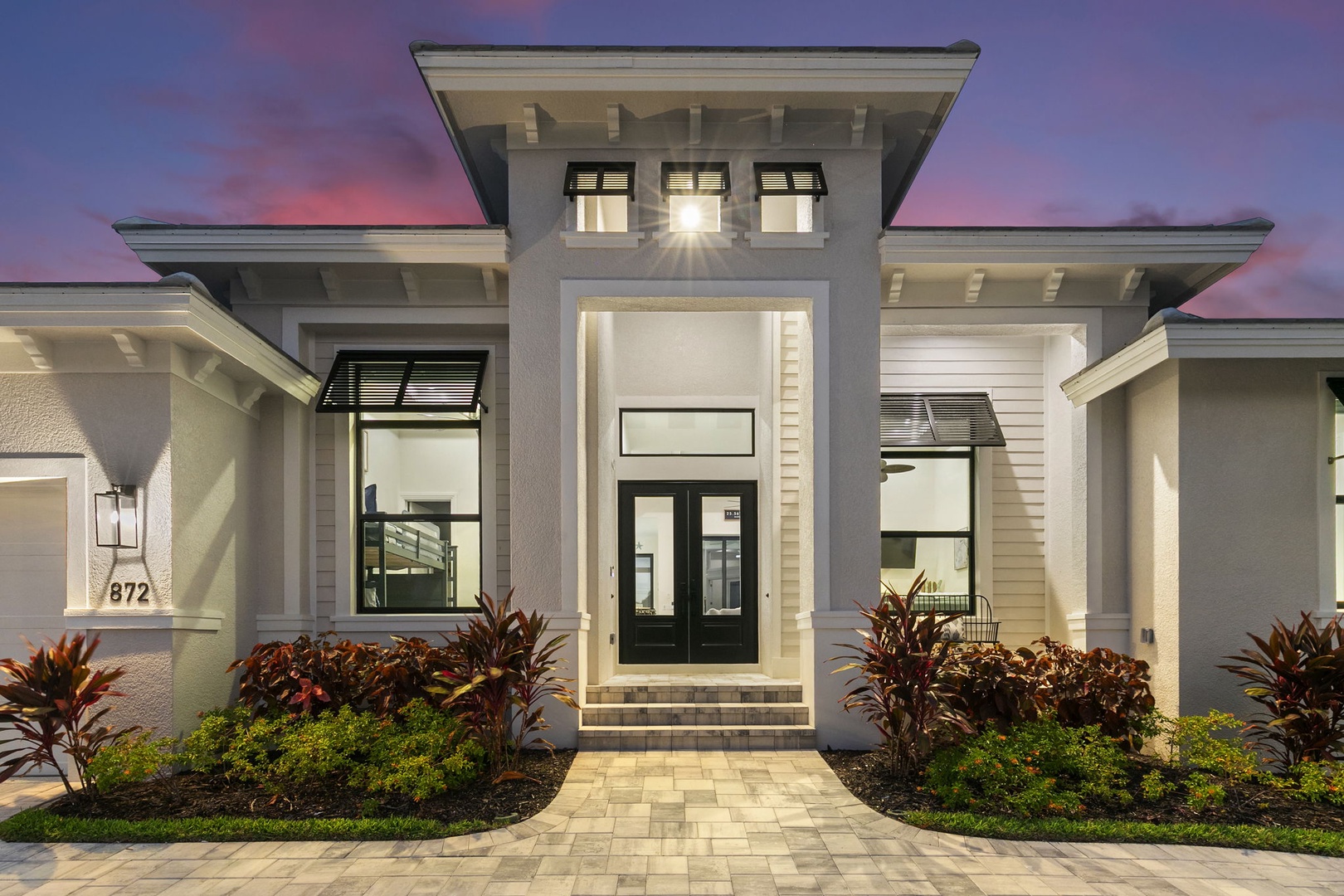 A stunning modern facade welcomes arrivals with clean architectural lines and warm lighting as evening descends. The property's entrance features contemporary design with dramatic roof details and manicured tropical landscaping.