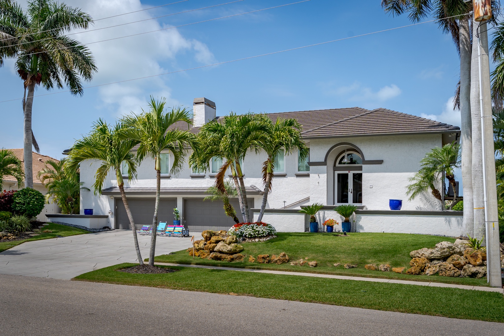 A spacious two-story home features white stucco exterior, Mediterranean-style architecture, and lush tropical landscaping with mature palm trees throughout the manicured lawn.