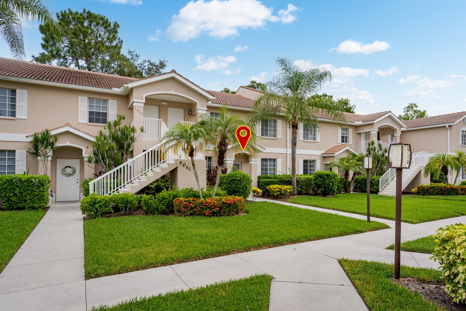 This Mediterranean-style property features exterior stairs and a well-maintained tropical landscape with lush lawns, palm trees, and vibrant flowering shrubs. A marked entrance clearly identifies the rental unit location.