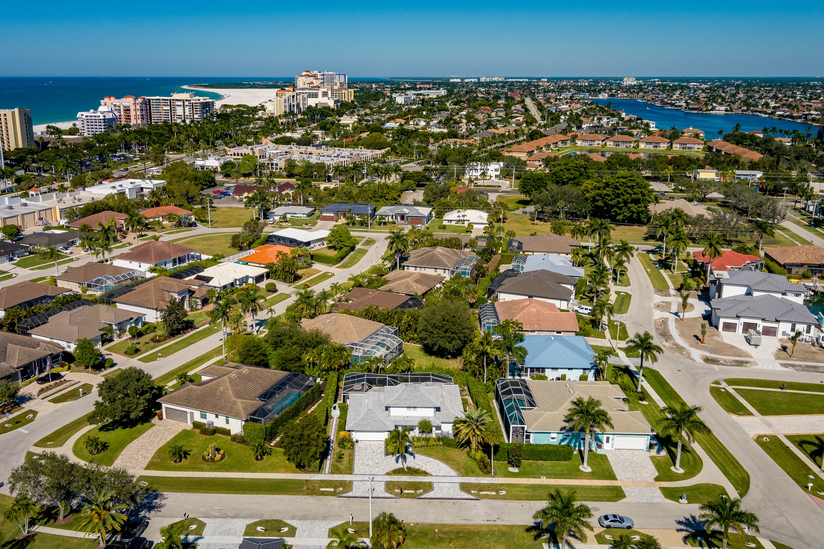 Aerial view of a coastal residential neighborhood featuring single-family homes with swimming pools, palm trees, and proximity to beaches and waterfront areas.