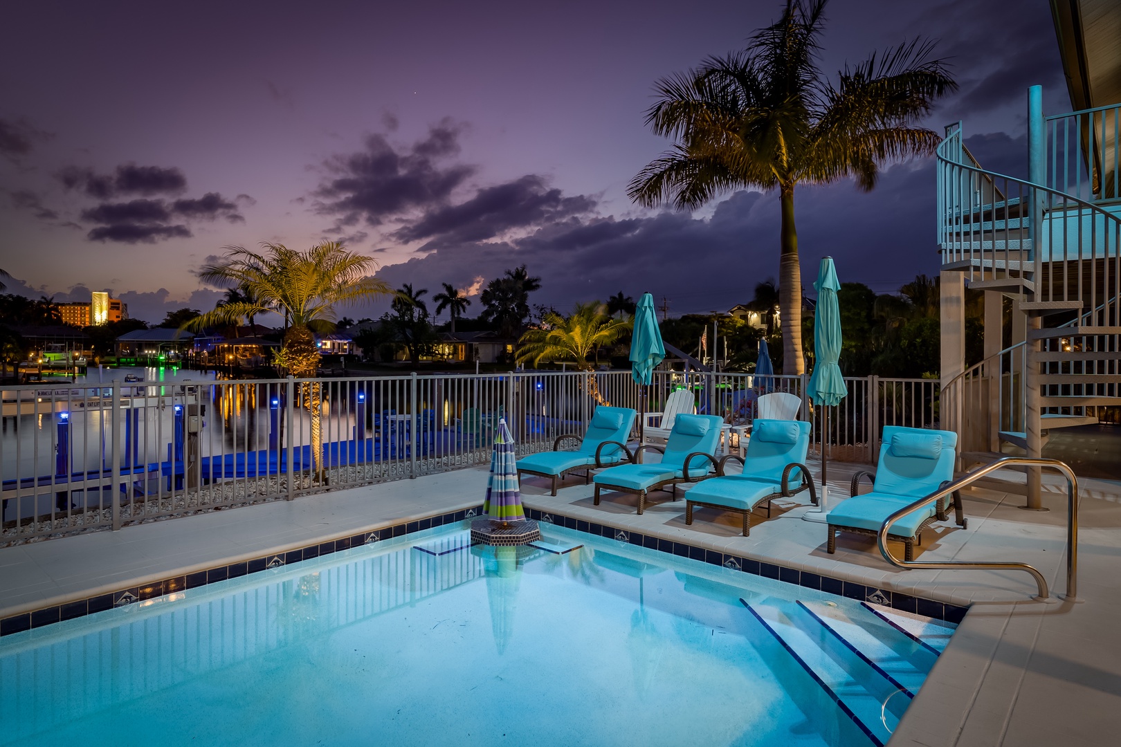 The property features a waterfront pool area with vibrant turquoise loungers and a scenic canal backdrop, framed by swaying palms against a dusky tropical sky.